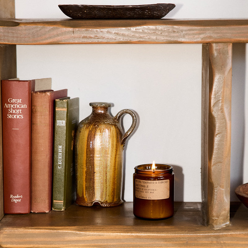 P.F. Candle Co. Standard Candle in Teakwood & Tobacco on a wood shelf with books and a vase